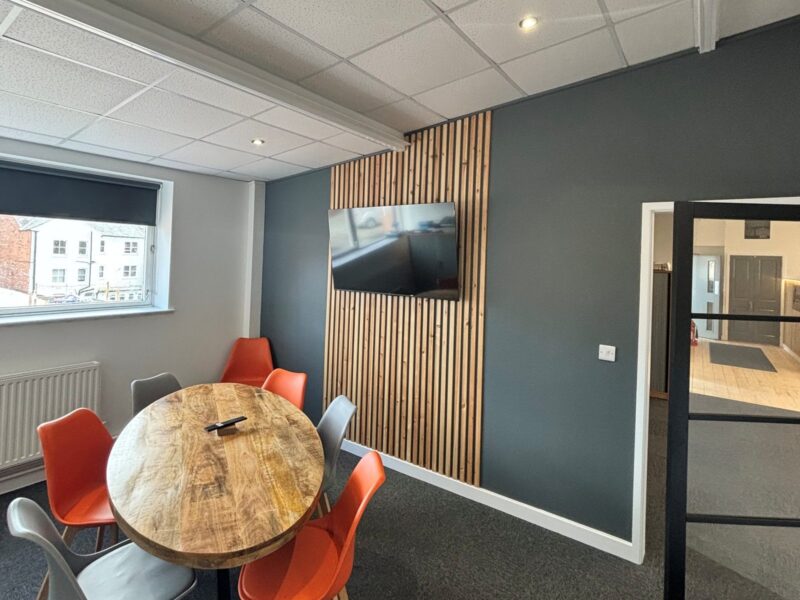 Meeting room with a round table and orange and grey chairs, beside a timber slat feature wall with a wall-mounted screen