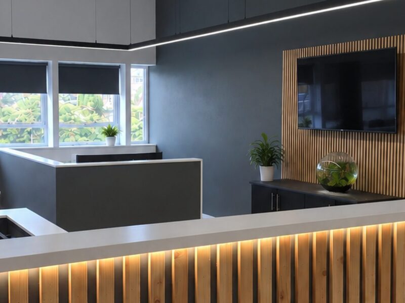 Completed office fit-out with a backlit timber slat reception counter, suspended linear lighting and a wall-mounted screen