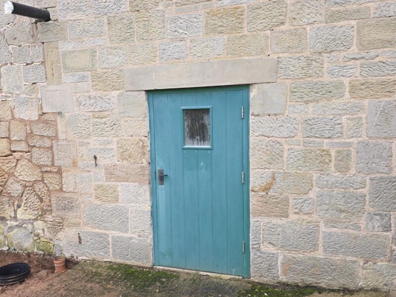 Weathered plank door painted teal, with a small glazed panel, set into the coursed stone elevation beneath a stone lintel