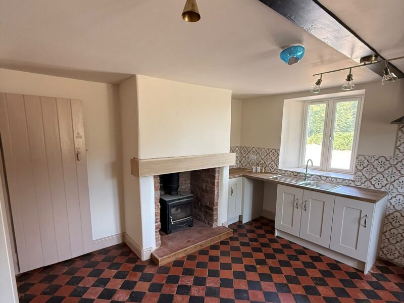 Living and kitchen area with a wood burning stove set into a brick fireplace and a red and black chequered tiled floor