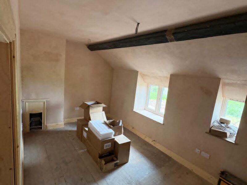 Bedroom plastered out beneath a dark beam, with a small fireplace in the far wall and boxes stacked on the bare floorboards