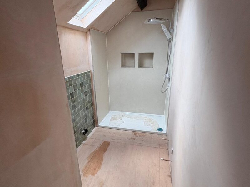 Shower room under construction, with the tray set in, the shower wall lined, the riser and head fitted and green tiling started alongside