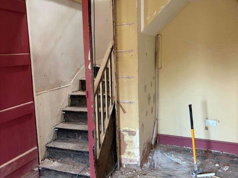 The original staircase before the works, with plaster stripped back from the walls, debris across the tiled floor and a sledgehammer standing in the room alongside