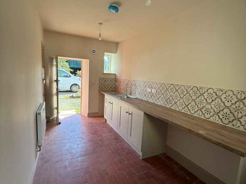Utility room with a run of units, a timber worktop, a patterned tiled splashback and a tiled floor, with the door open to outside